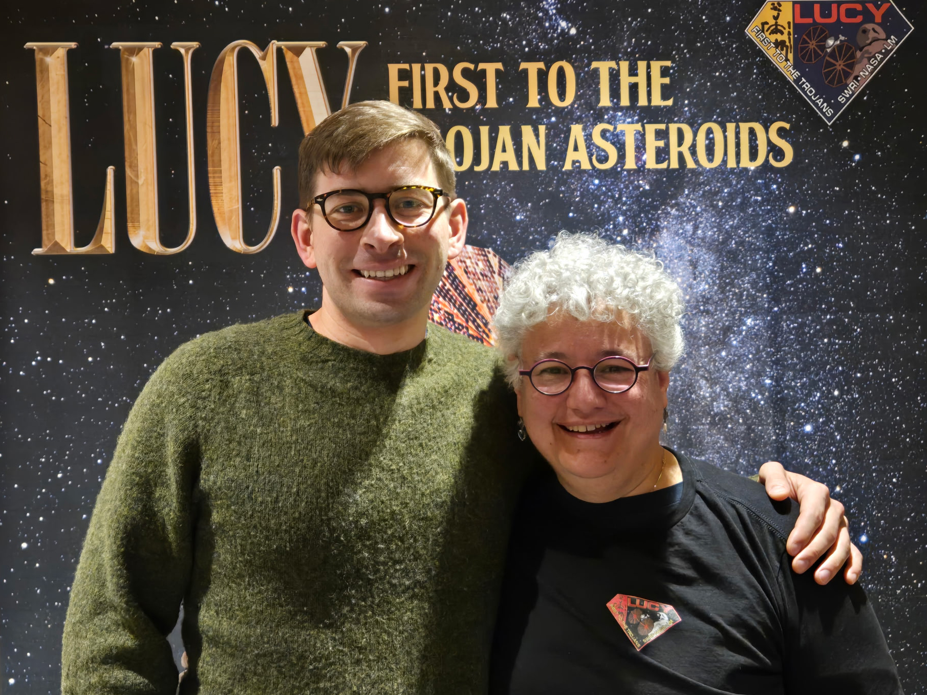 Two people - Harrison Agrusa and Jess Sunshine - posing in front of banner for Lucy Mission