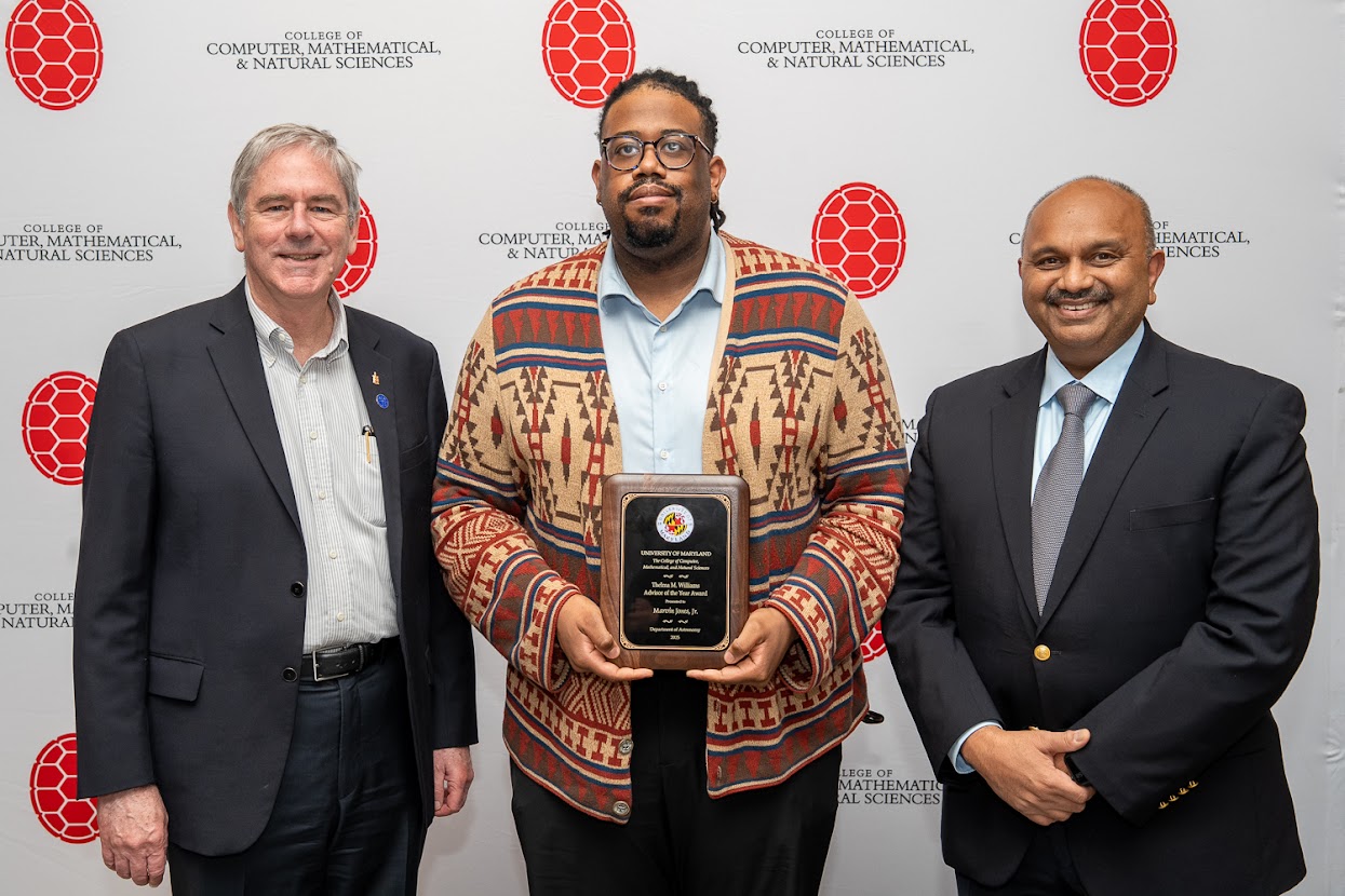 Three men standing in front of a CMNS-themed backdrop. Astronomy-chair Dr. Andrew Harris on the left, award recipient Marvin Jones in the middle holding his placque, and CMNS Dean Amitabh Varshney on the right.